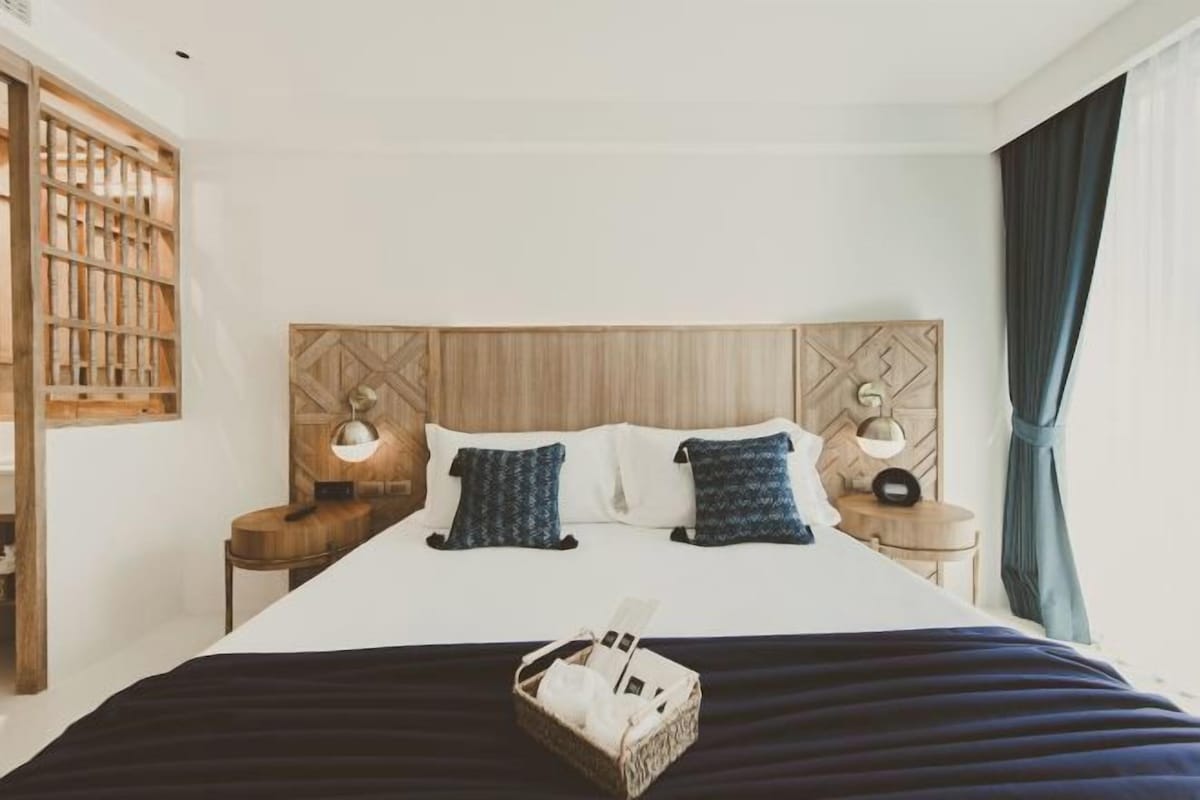 A hotel room with a bed, two bedside tables, a headboard, and a window with curtains.