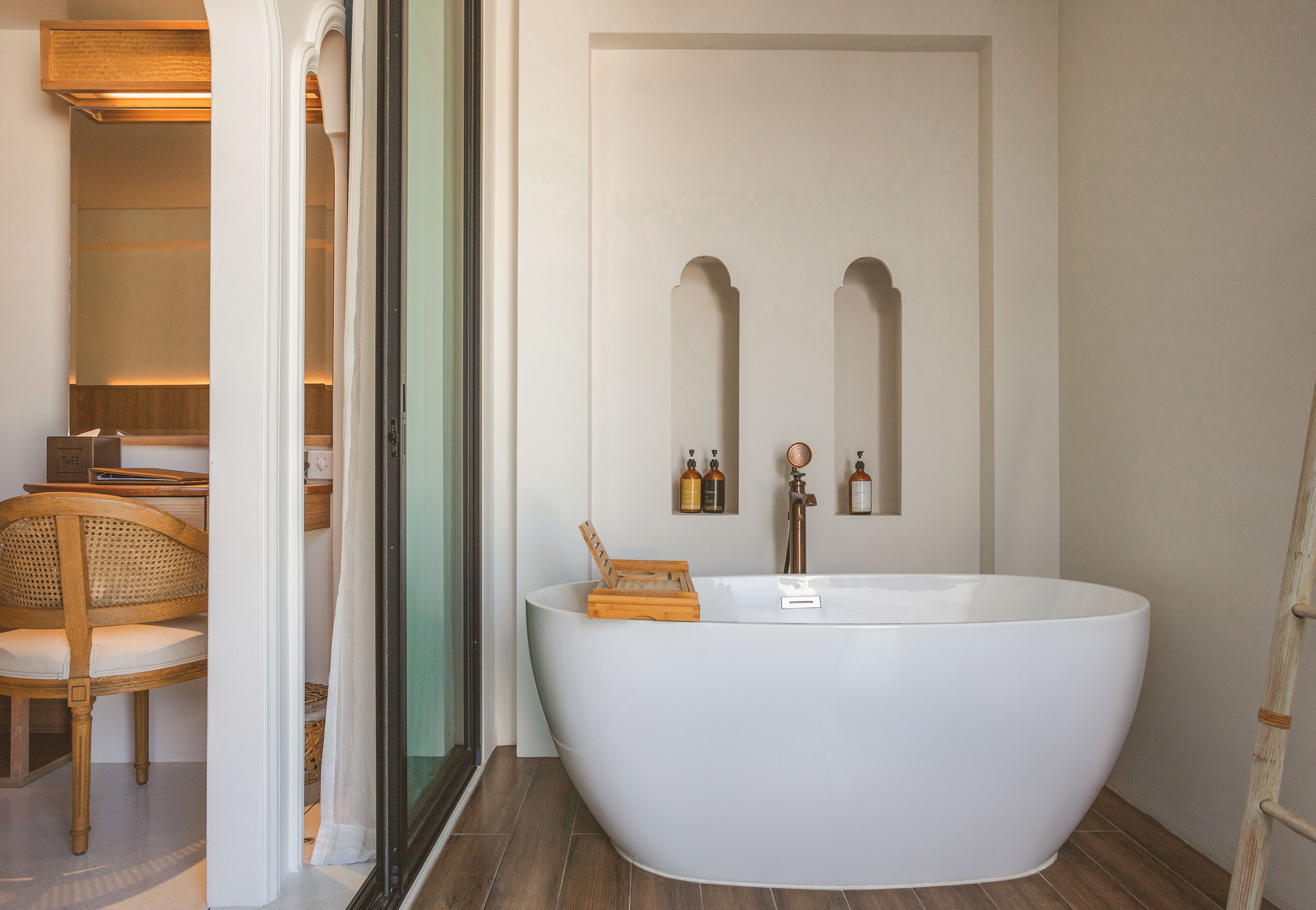 A modern bathroom with a freestanding bathtub, wooden chair, and a mirror with two bottles of lotion.