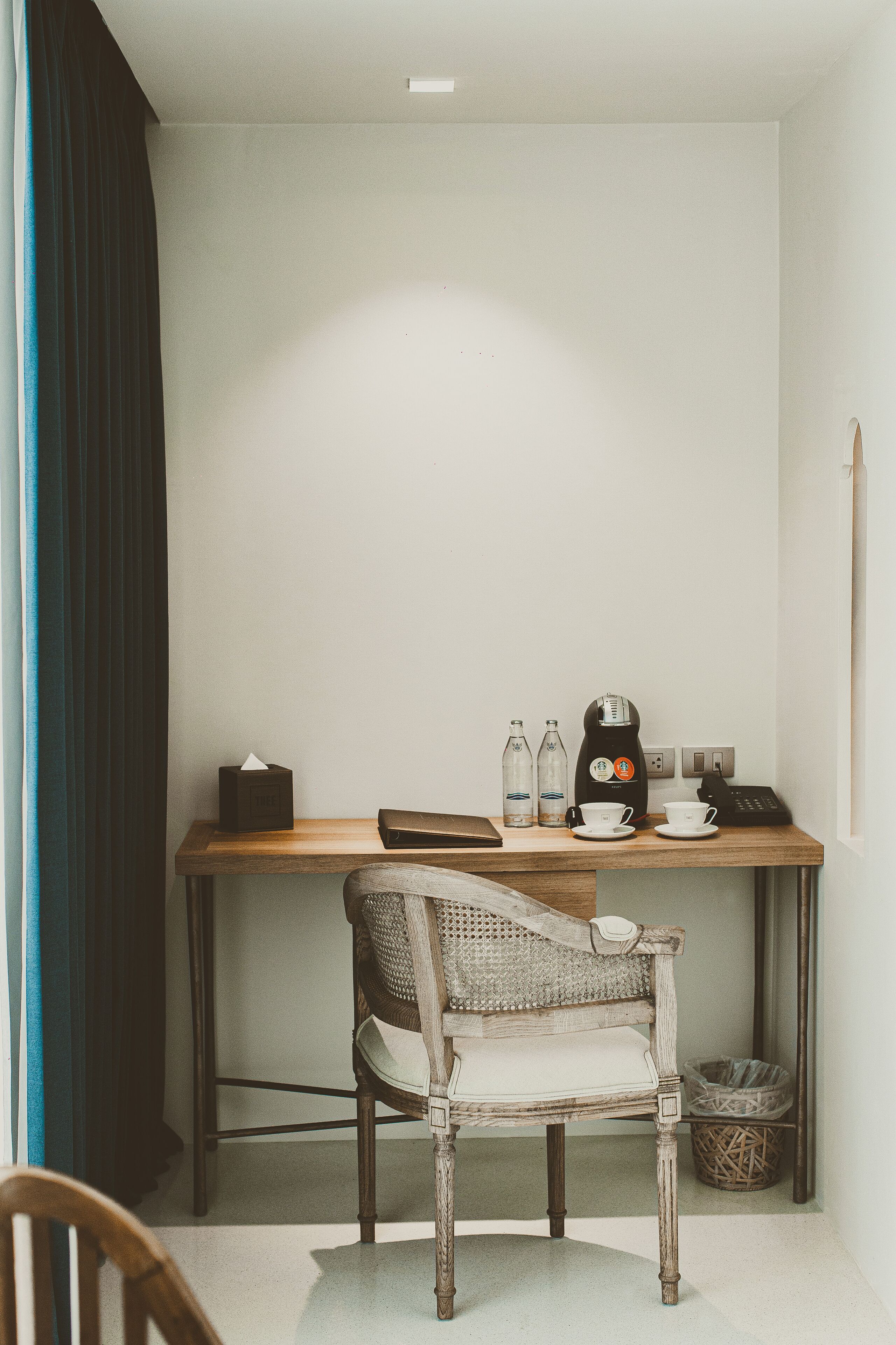 A small, simple room with a wooden desk, a wicker chair, a trash bin, and a wall-mounted telephone.