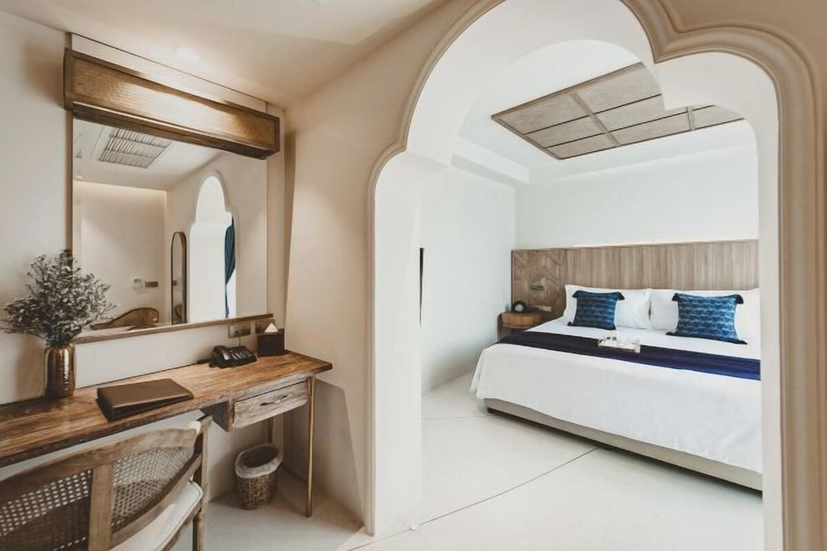 A bedroom with a bed, a desk with a chair, a mirror, and a small plant.