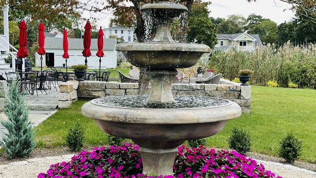 Una fuente de piedra de varios niveles rodeada de flores rosas vibrantes, con sombrillas rojas y mesas al aire libre al fondo.
