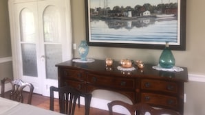 Un área de comedor con una mesa de madera, sillas, una puerta blanca con paneles de vidrio y un cuadro enmarcado que muestra una escena portuaria en la pared.