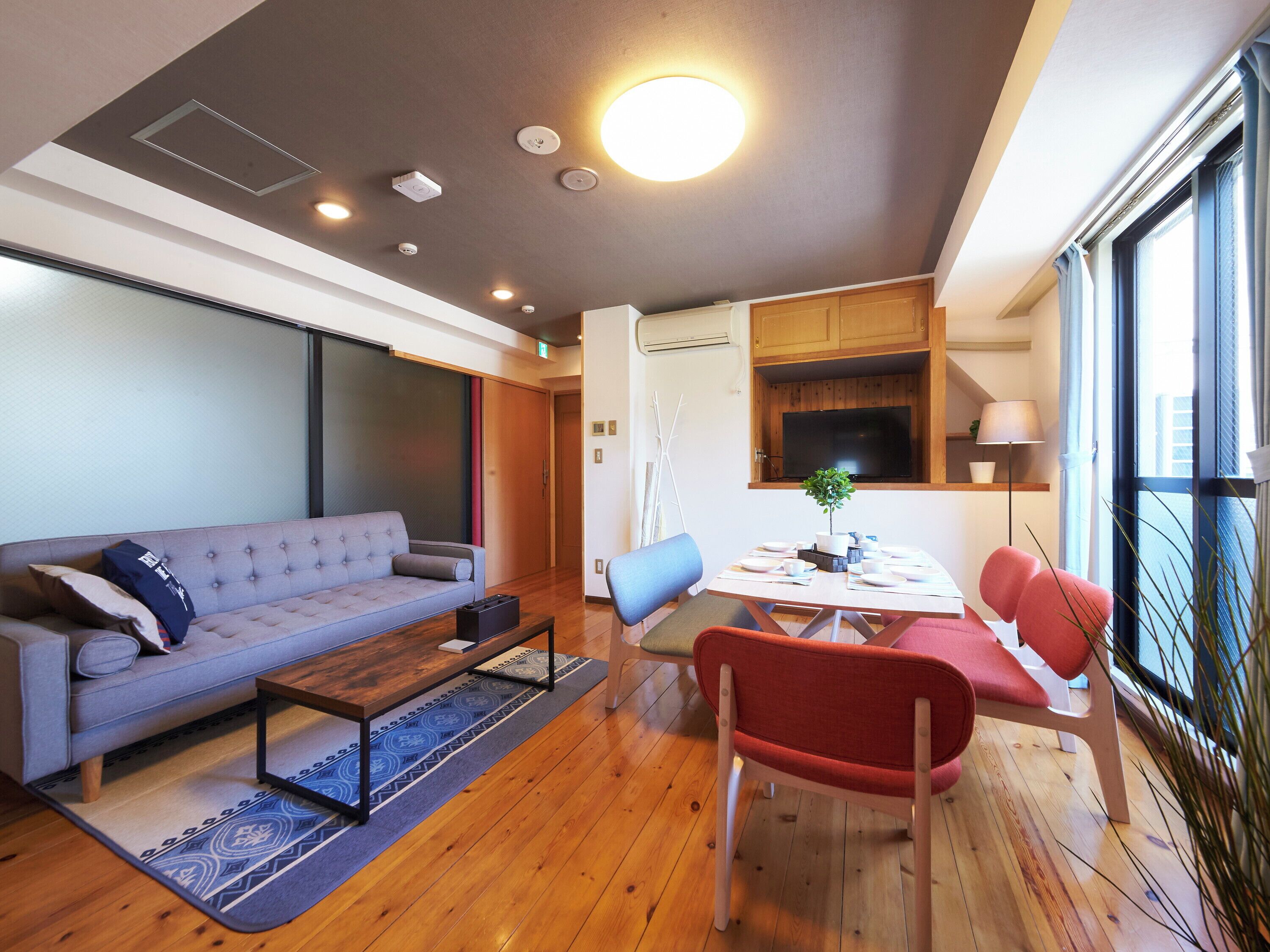 A modern living room with a grey sofa, a wooden coffee table, and a dining table with chairs. There is a TV mounted on the wall, a plant on the table, and a window with curtains.