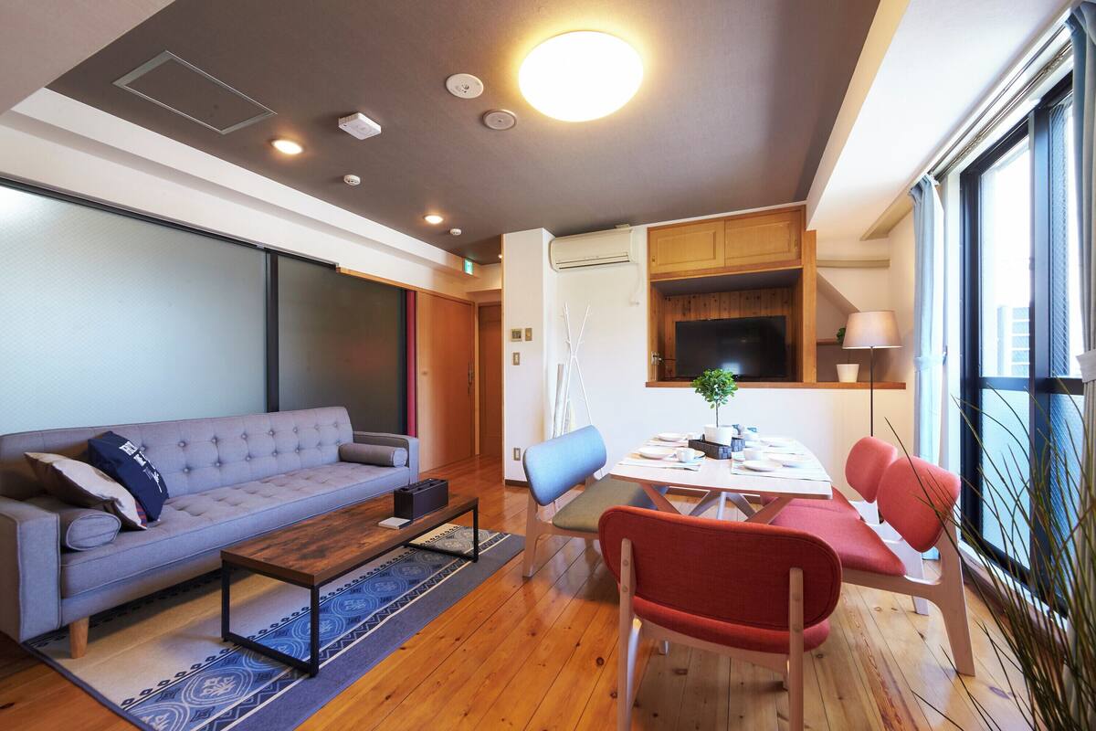 Una sala de estar moderna con un sofá gris, una mesa de centro de madera y una mesa de comedor con sillas. Hay un televisor montado en la pared, una planta sobre la mesa y una ventana con cortinas.