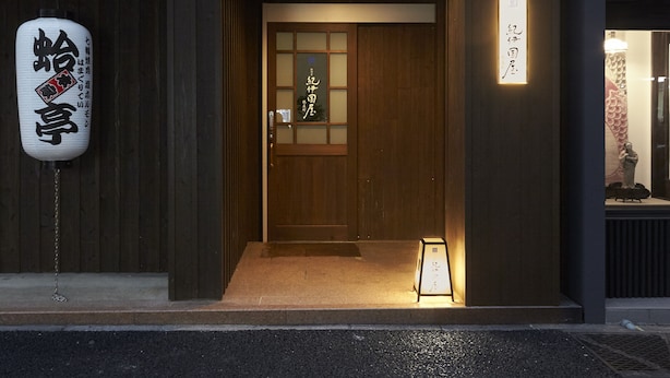Une entrée de bâtiment japonais traditionnelle, avec une porte en bois, une enseigne avec des caractères japonais et un petit panneau lumineux à l’extérieur.