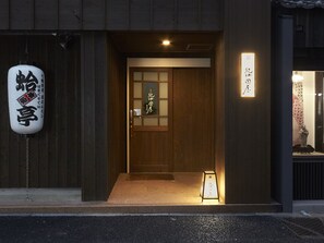 A traditional Japanese building entrance with a wooden door, a sign with Japanese characters, and a small illuminated signboard outside.