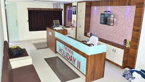 A hotel reception area with a wooden reception desk, a television mounted on the wall, and a large window with curtains. - The New Holiday Inn (Mahesana)
