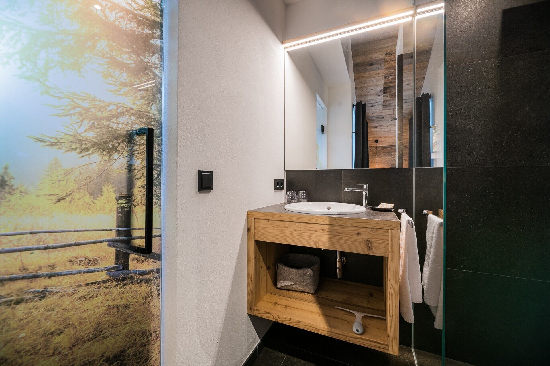 Un baño moderno con tocador de madera, un espejo grande y vista a un bosque a través de una puerta de vidrio.