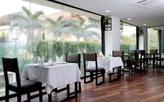 Un restaurante con mesas dispuestas para comer, amplias ventanas con vistas a palmeras y edificios, y suelo de madera.