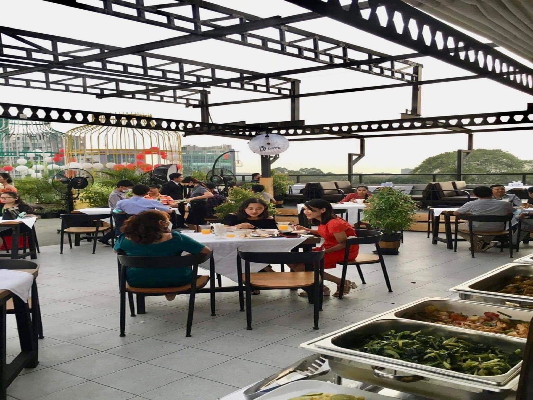 Un área de comedor al aire libre con mesas y sillas, rodeada por una estructura metálica y plantas en macetas.