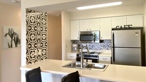 A modern kitchen with white cabinets, a stainless steel refrigerator, and a microwave. There are two black chairs at the kitchen counter. - Pointe Royale Condo retreat (Branson)