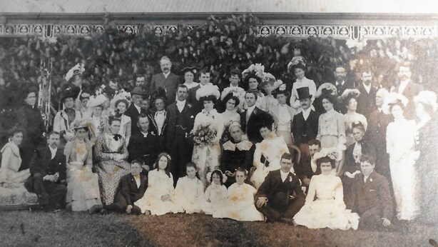 Ett historiskt svartvitt fotografi som föreställer en stor grupp människor i formella kläder, samlade utomhus med ett dekorativt räcke i bakgrunden.