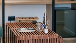 Una habitación de hotel moderna con una mesa de centro de madera, una cama con ropa de cama blanca, una mesita de noche con lámpara, un refrigerador pequeño y un estante de pared con hervidor y tazas.
