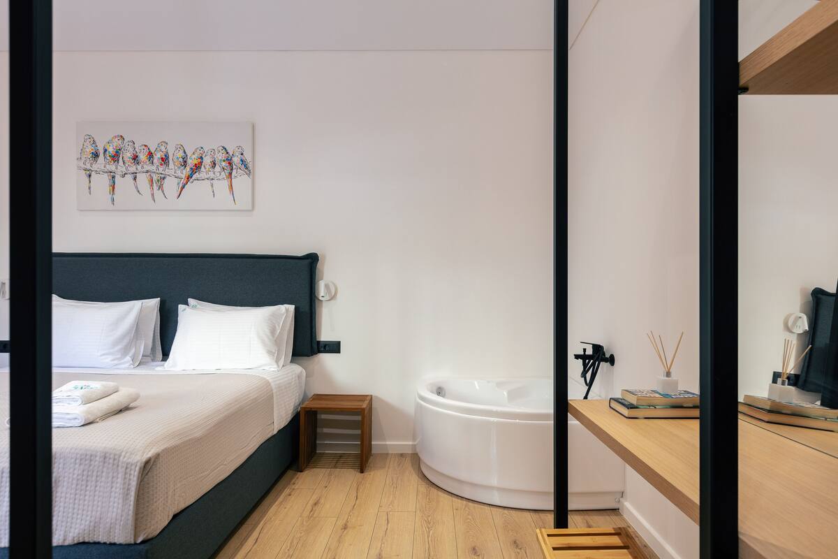A modern bedroom with a bed, a small bedside table, a nightstand, a wall-mounted artwork, a white bathtub, and a wooden shelf with books and a lamp.