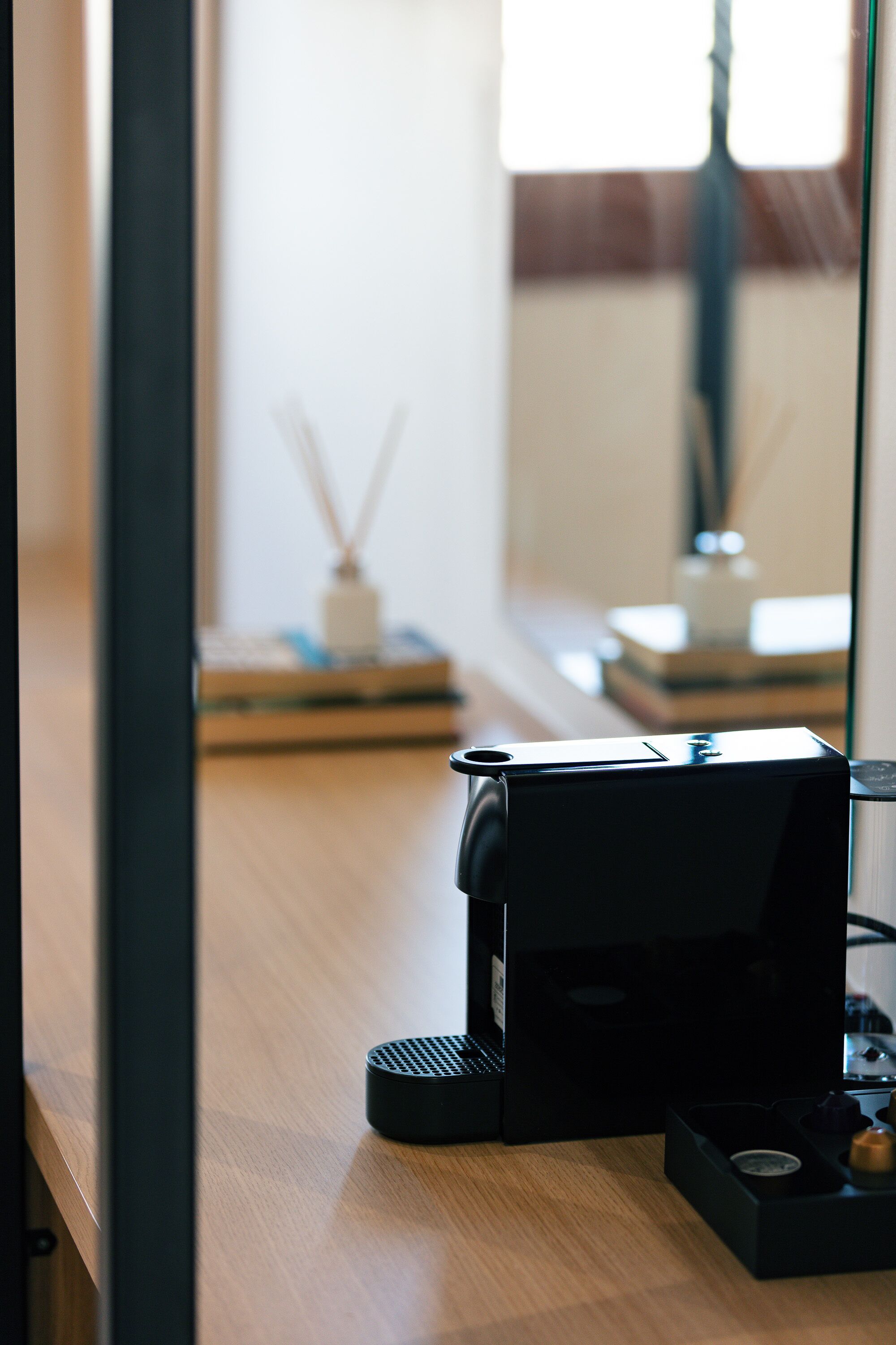 Une machine à café est posée sur une table en bois. Un miroir reflète une fenêtre et une pièce dont le mur est blanc, et on y aperçoit un petit objet décoratif.