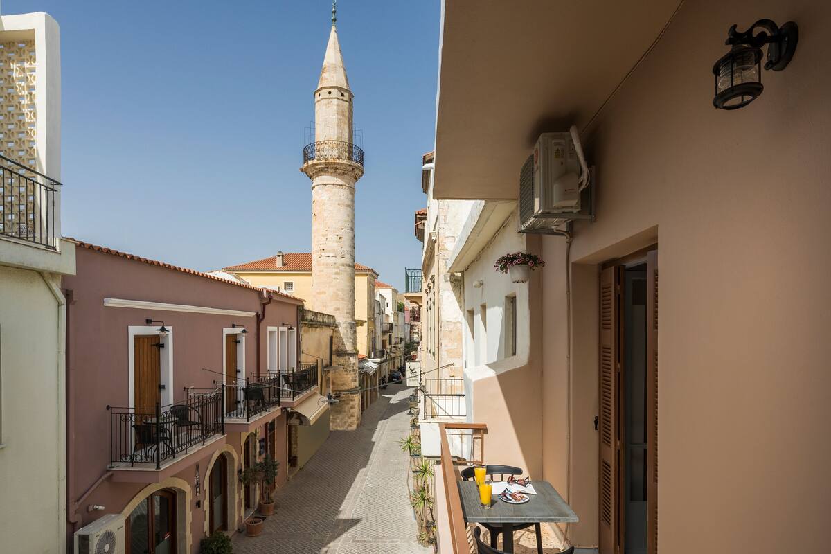 A narrow street with buildings on either side, a prominent tower, and a table with a drink on a balcony.
