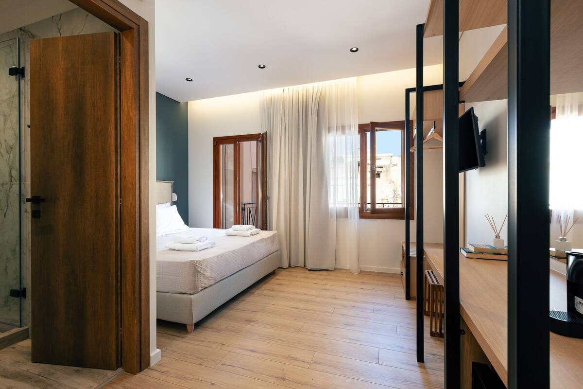 A modern bedroom with a wooden floor, a bed with white linens, a bedside table, a TV, and a window with curtains.