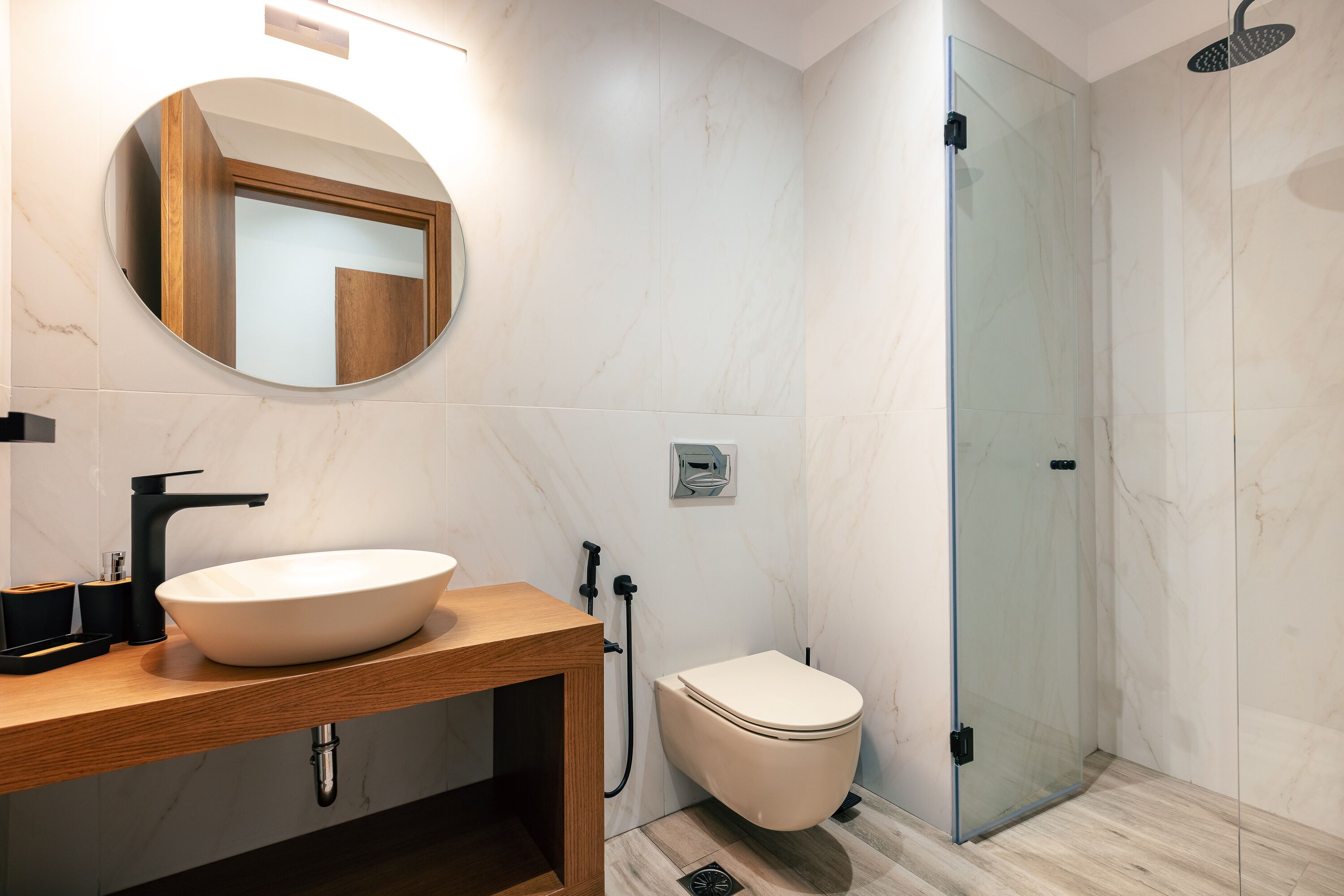 A modern bathroom with a round mirror, a wooden vanity, a white sink, a toilet, and a glass-enclosed shower area.