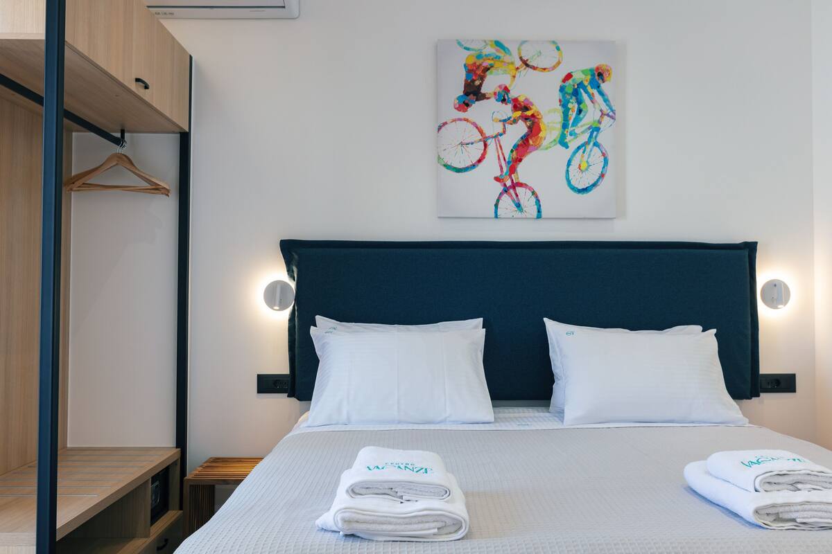 A hotel room with a bed, white pillows, a headboard, and a colorful bicycle artwork on the wall.