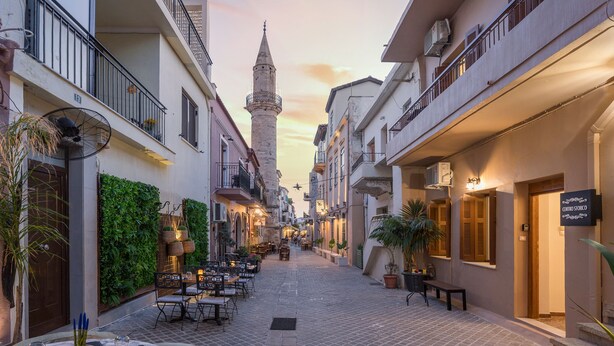 Una calle estrecha con mesas al aire libre, una torre al fondo y edificios con balcones.
