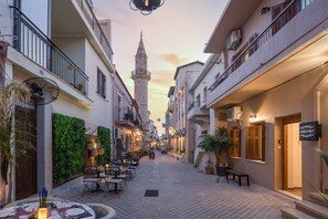Une rue étroite avec des tables en terrasse, une tour en arrière-plan et des bâtiments dotés de balcons.