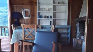 Een houten blokhut van binnen, met een eethoek, een blauwe stoel, een boekenkast en een stenen open haard.