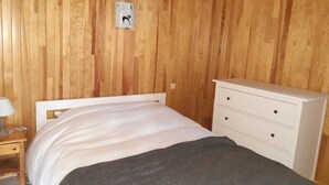 Een slaapkamer met een houten lambrisering, een wit bed met een grijze deken, en een witte ladekast met drie lades.