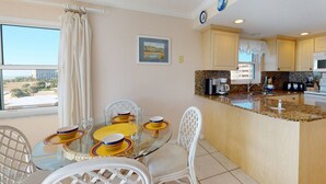 A modern kitchen with a dining area, featuring a glass table, white chairs, and a view of the cityscape through large windows. - Fabulous Location. Updated Beachfront Condo. Pool, Recreation Amenities, Comfortable! (Treasure Island)