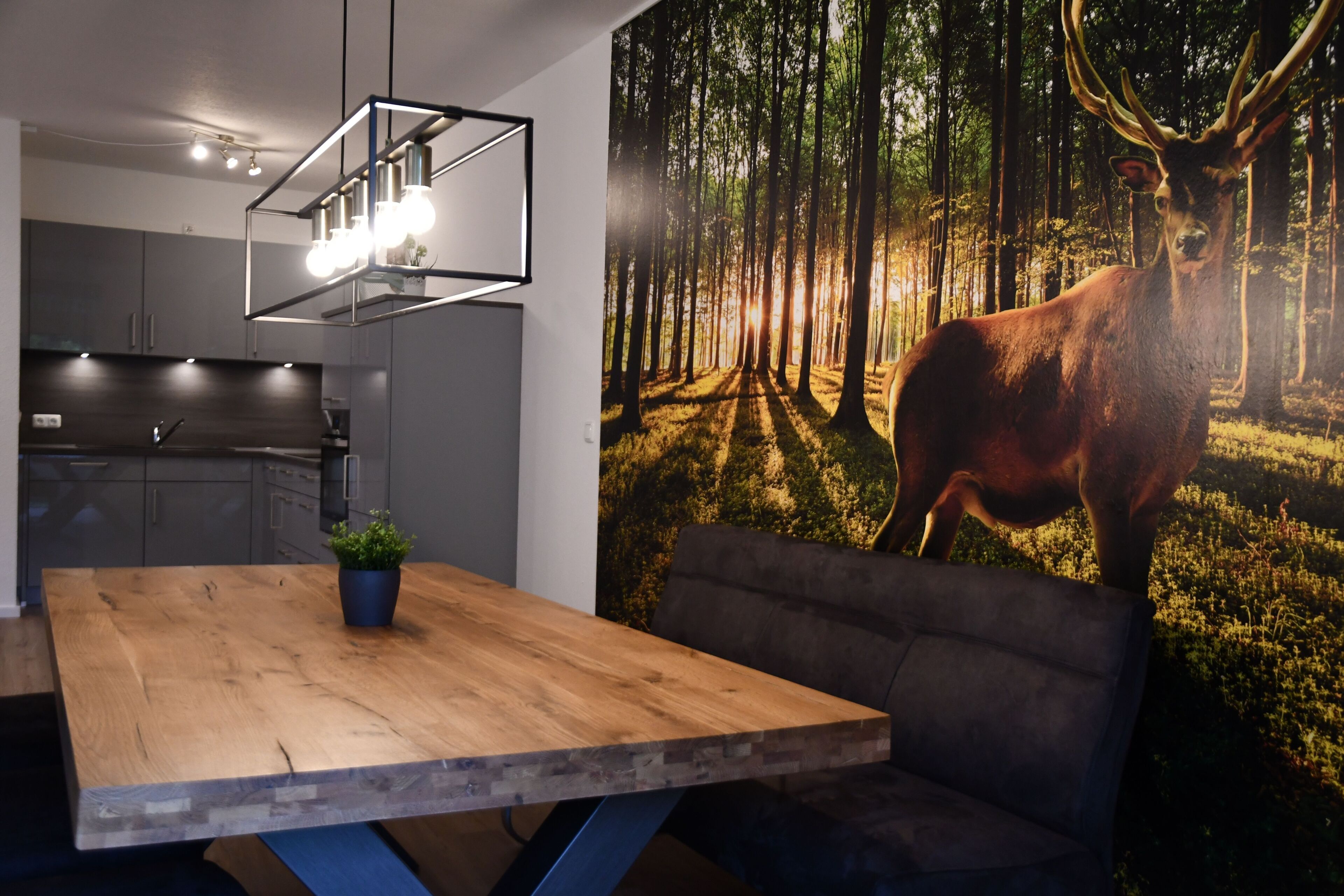 Eine moderne Küche mit grauen Schränken, einem Holztisch mit einer Pflanze und einem Wandbild mit Waldmotiven.
