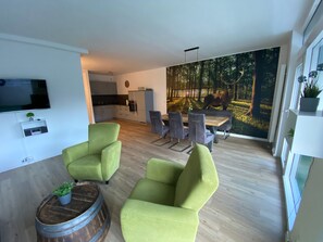Ein modernes Wohnzimmer mit grünen Sesseln, einem Holztisch und einem Essbereich mit einem großformatigen Wandbild von einem Wald.