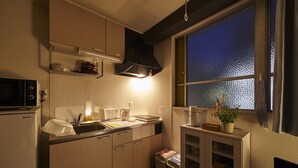 A compact kitchen with a microwave, sink, and cabinets. A window with a view of the water is visible. - Oyado Kinokuniya Daikoku Machi (Nagasaki)