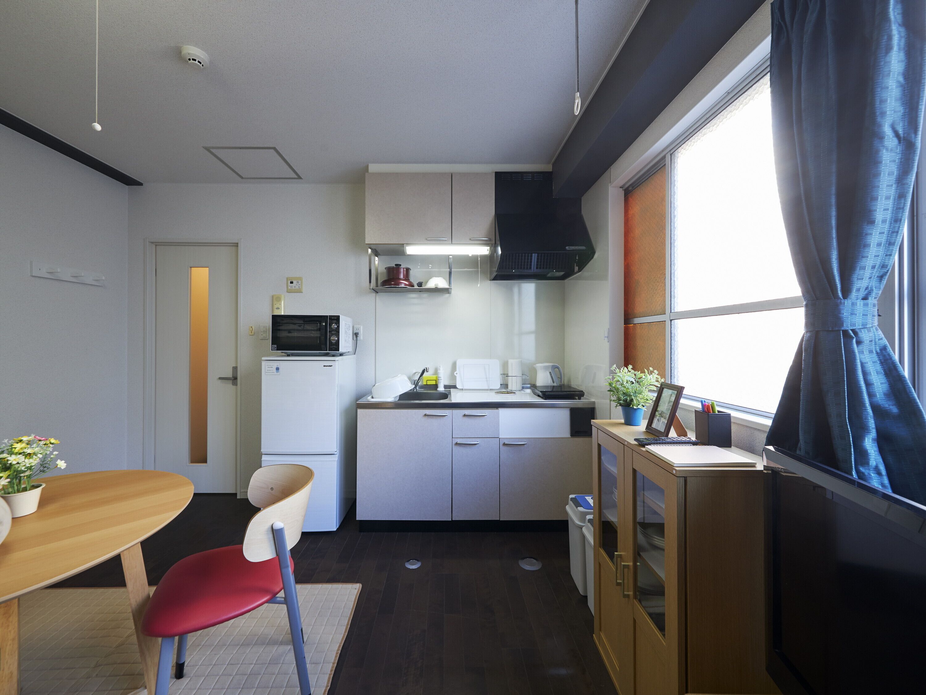 A compact kitchen with a microwave, stove, and sink. There is a dining table with chairs, a small cabinet, and a window with curtains.