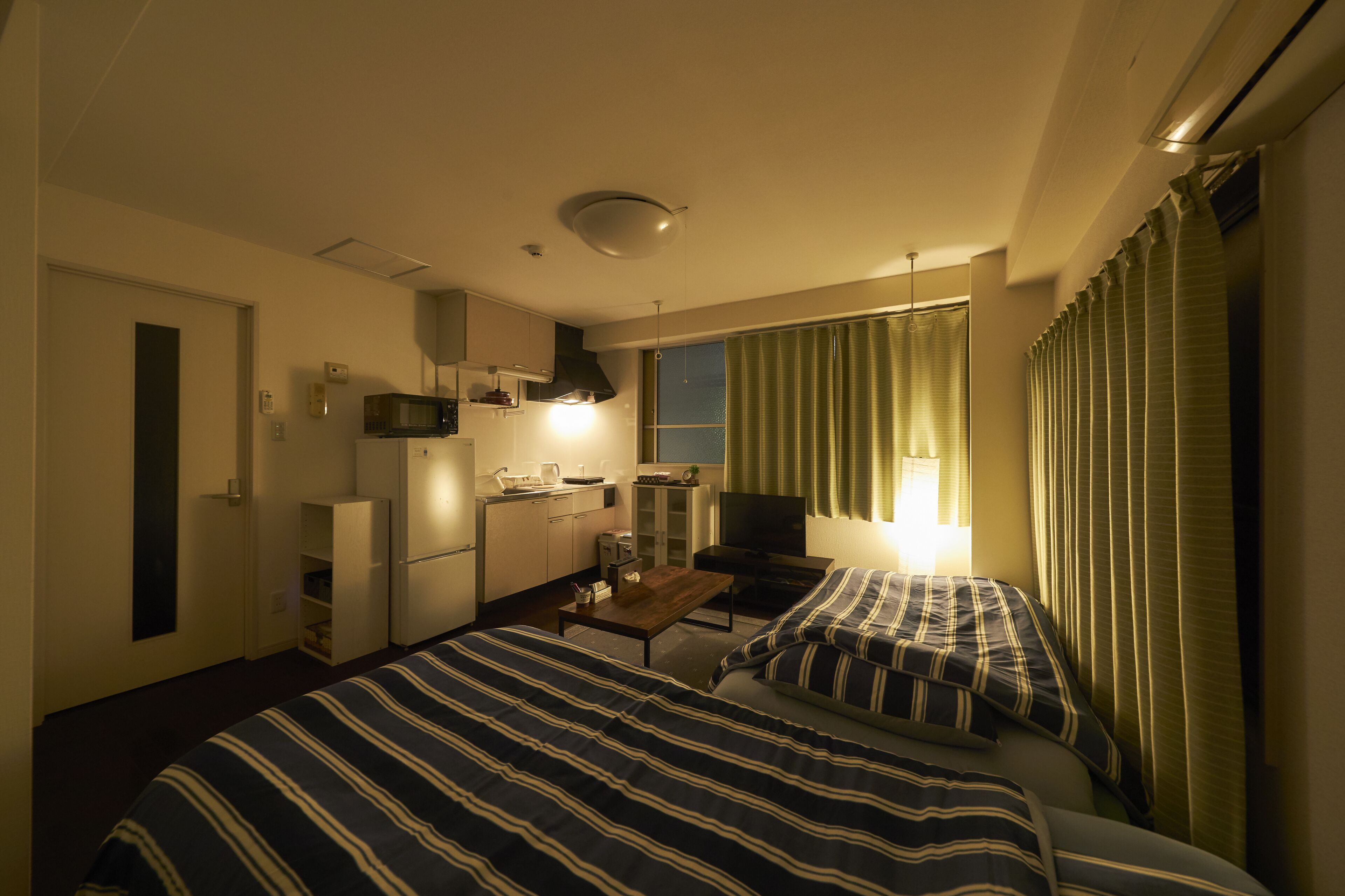 Quarto de hotel com duas camas, uma kitchenette, televisão e janela com cortinas.