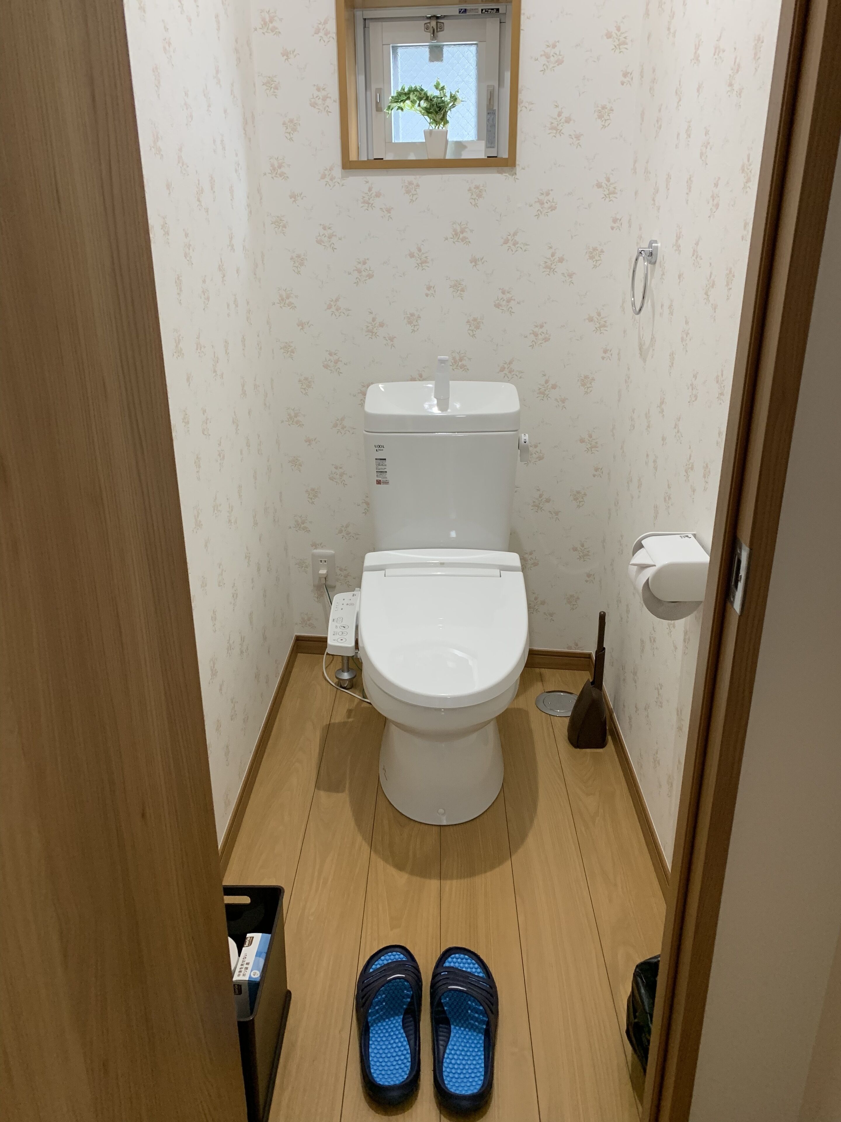 Un baño pequeño con inodoro, una ventana con una planta y un par de zapatillas en el suelo.
