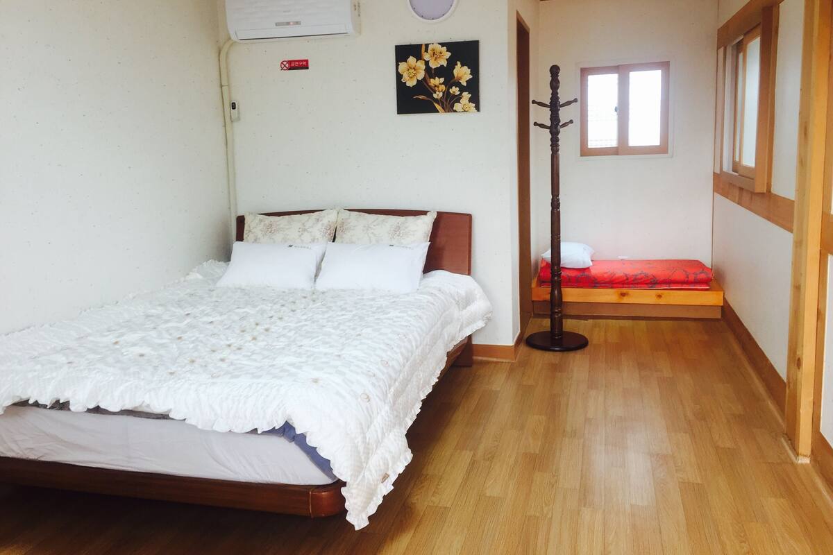 A bedroom with a wooden floor, a bed with white bedding, a small bedside table, a wall-mounted air conditioner, a clock, and a window with a view of the outside.