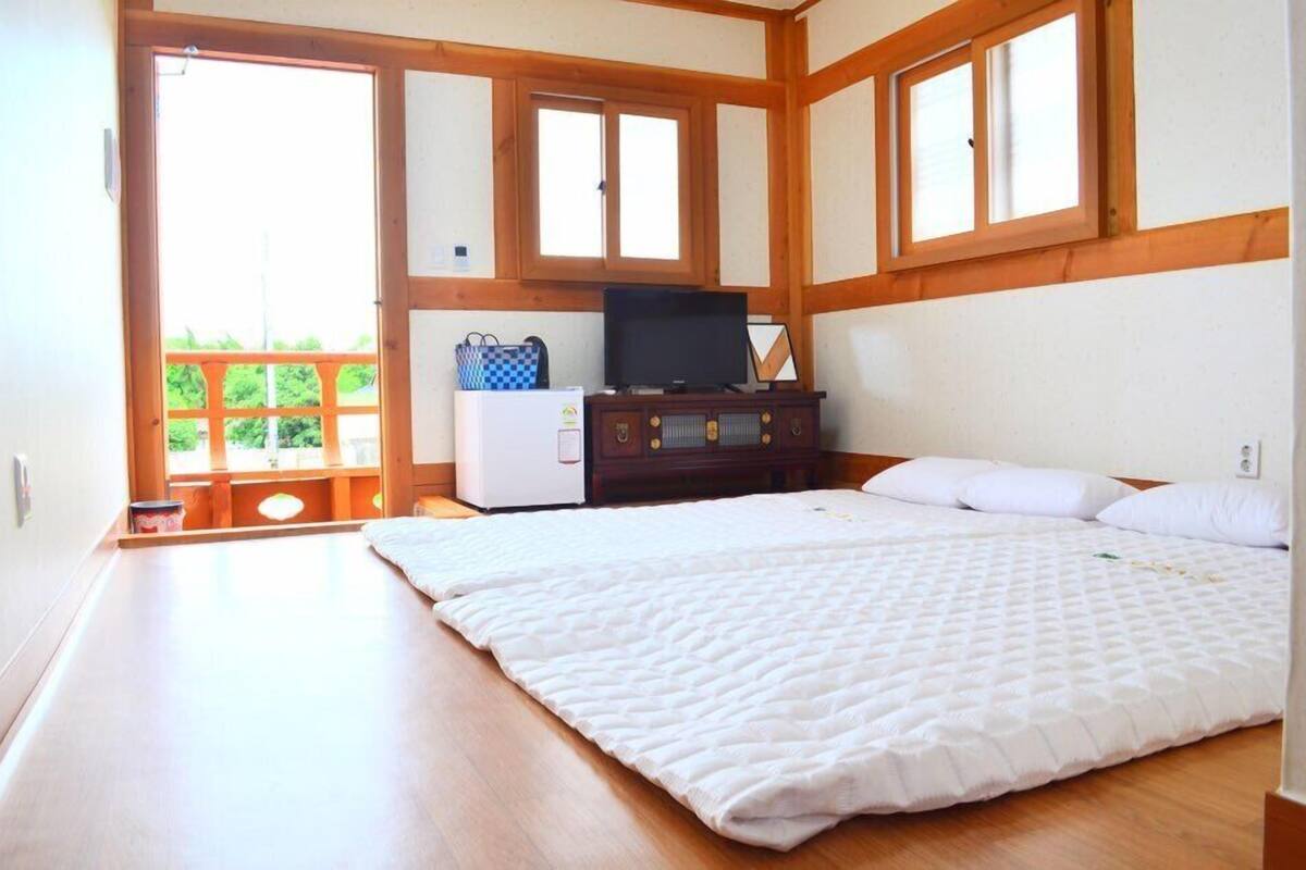 A bedroom with a large bed, wooden ceiling beams, a television on a dresser, and a balcony with a view of greenery.