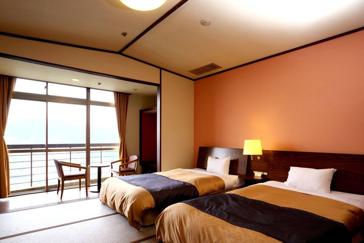 Une chambre d’hôtel avec deux lits, un coin salon comprenant une chaise et une table, et une grande fenêtre donnant sur un paysage agréable.