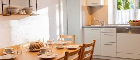Una cocina con armarios blancos, una mesa de comedor de madera y sillas. Se ve vegetación a través de una ventana.