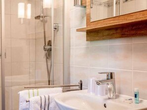 A bathroom with a glass shower enclosure, a white sink with a chrome faucet, and a wooden shelf above the sink.
