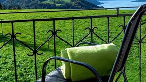 Una silla con cojín verde en un balcón con barandilla metálica, con vistas a un paisaje verde y una masa de agua.