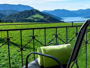 Una silla con cojín verde en un balcón con barandilla metálica, con vistas a un paisaje verde y una masa de agua.