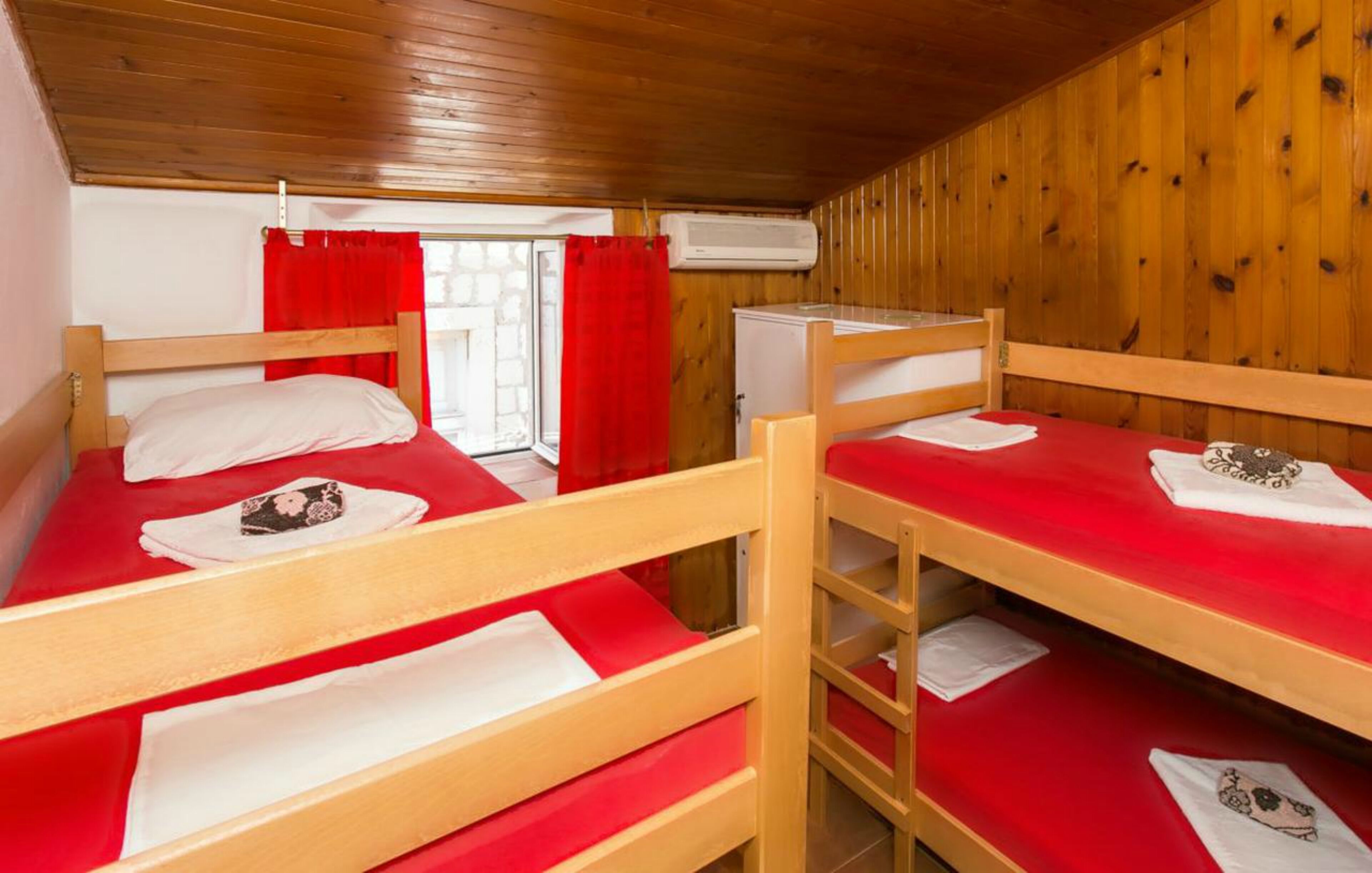 A cabin with wooden walls and floors, two bunk beds with red bedding, a small window with red curtains, and an air conditioning unit.