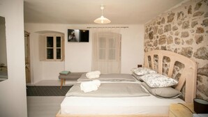 A bedroom with a stone wall, a bed with pillows and a blanket, a small table, a TV on the wall, and a door with a curtain. - Rooms Epidaurus (Dubrovnik)