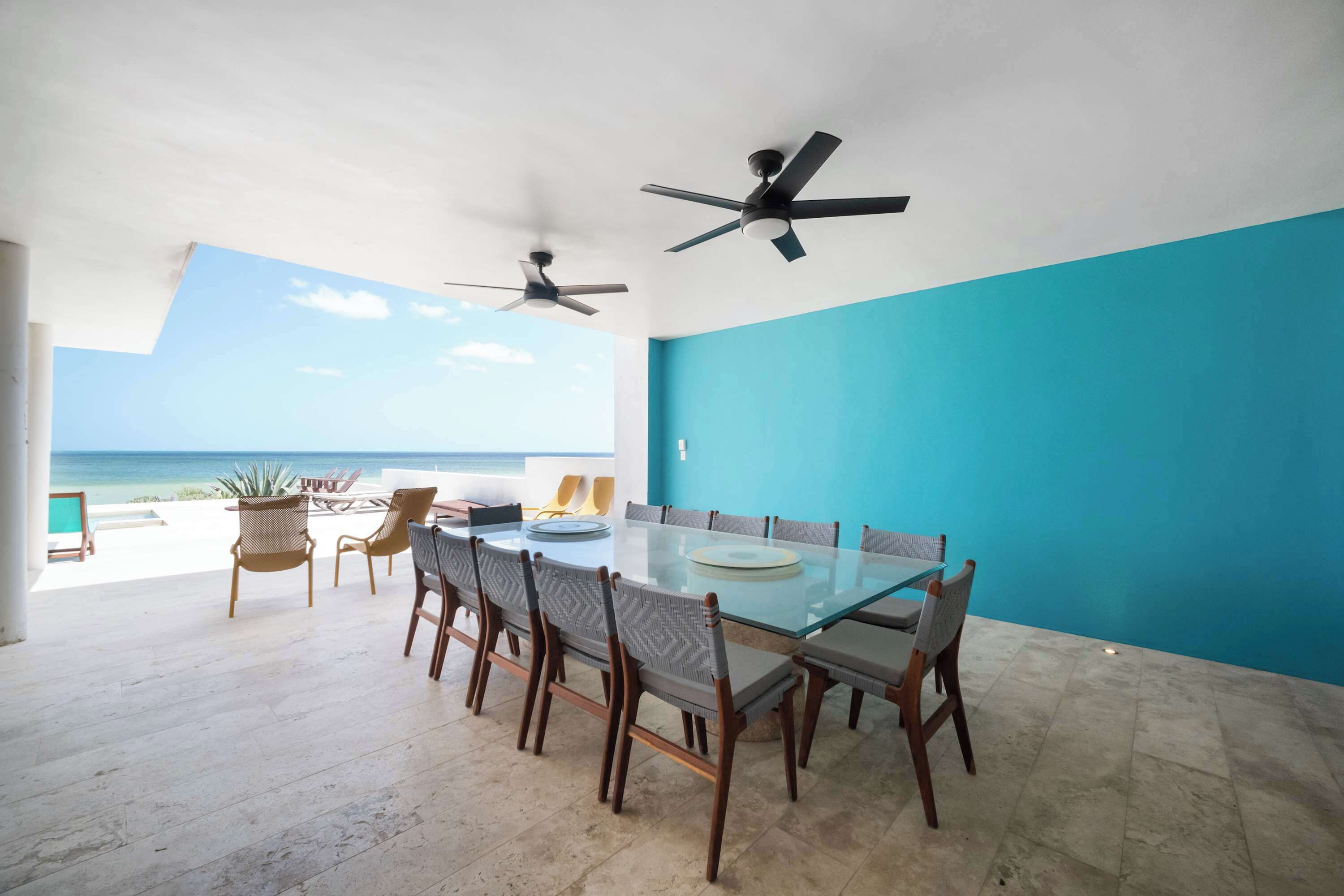 Comedor con mesa y sillas de vidrio, dos ventiladores de techo, vista al océano y una pared de acento azul.