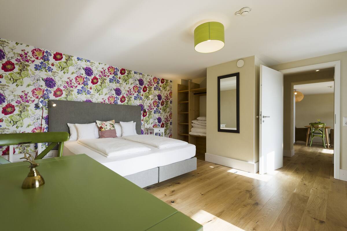 Un dormitorio con una cama, una mesa verde, un papel tapiz floral y un pasillo que lleva a un baño.