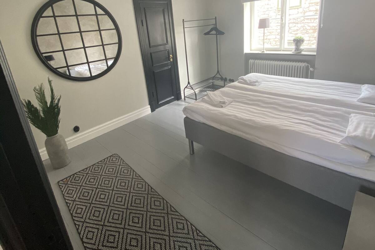A bedroom with a bed, a round mirror, a door, a radiator, and a window with a view of a stone building.