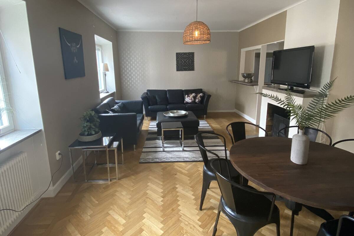 A spacious living area with a herringbone wooden floor, a dark-colored sofa, a small coffee table, and a dining table with chairs.
