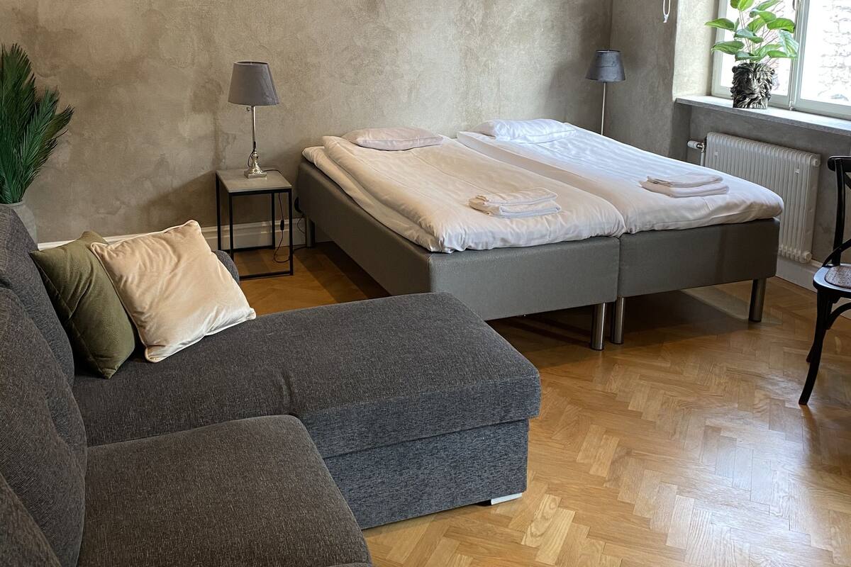A bedroom with a bed, a grey sofa, a small table, a lamp, and a window with a view.