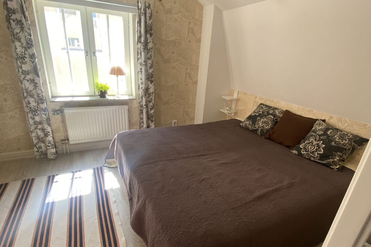 A bedroom with a large bed, a window with curtains, a radiator, and a striped rug.