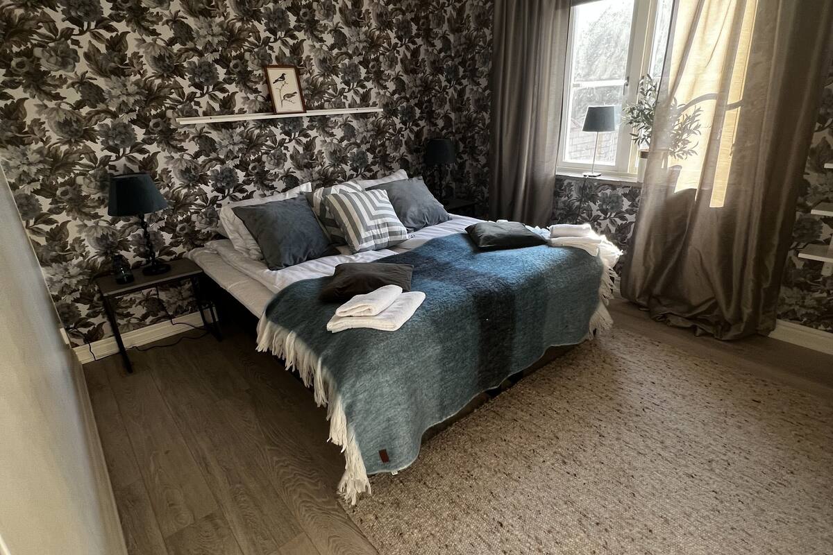 A bedroom with a bed, floral wallpaper, a ceiling light, a window with curtains, and a bedside table with a lamp.